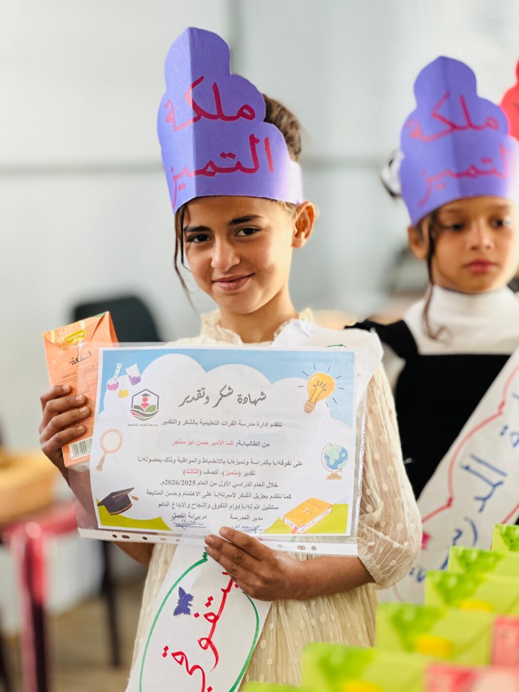 A girl wearing a Queen of Excellence crown holding her appreciation certificate from Al-Furat School in Deir al-Balah, Gaza
